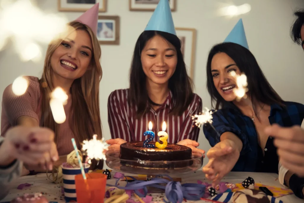 Três amigas comemoram aniversário de 23 anos com bolo, velas, chapéus de festa e muita alegria.